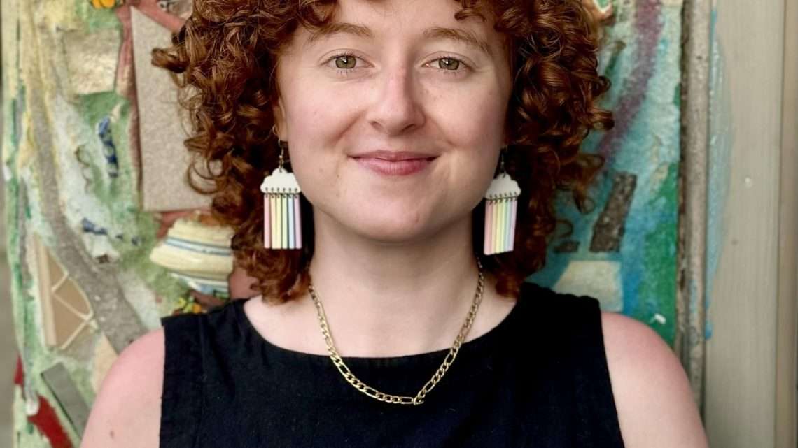 Bridget is standing in front of a multi-colored mosaic made by Philadelphia artist Isaiah Zagar. Their eyes are green, their skin is white, and their hair is red and curly. Their hair is just above shoulder length. They are smiling softly towards the camera. They are wearing a black sleeveless shirt, a gold chain necklace, and cloud earrings with rainbow colored streaks dangling off the clouds.