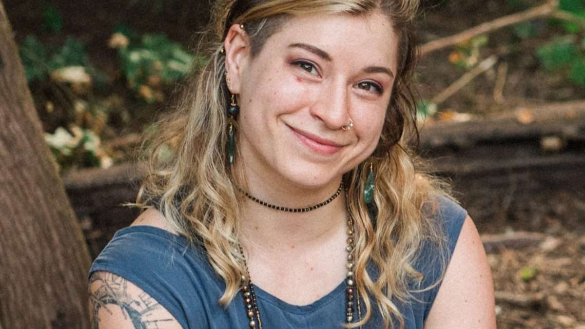 Gabrielle, a white person with blonde, wavy hair and hazel eyes, smiles at the camera and is surrounded by greenery and trees.