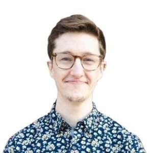 Image of Kaleb, a white person with short brown hair and glasses. Dressed in a dark blue button down with a small flower pattern, they smile at the camera.